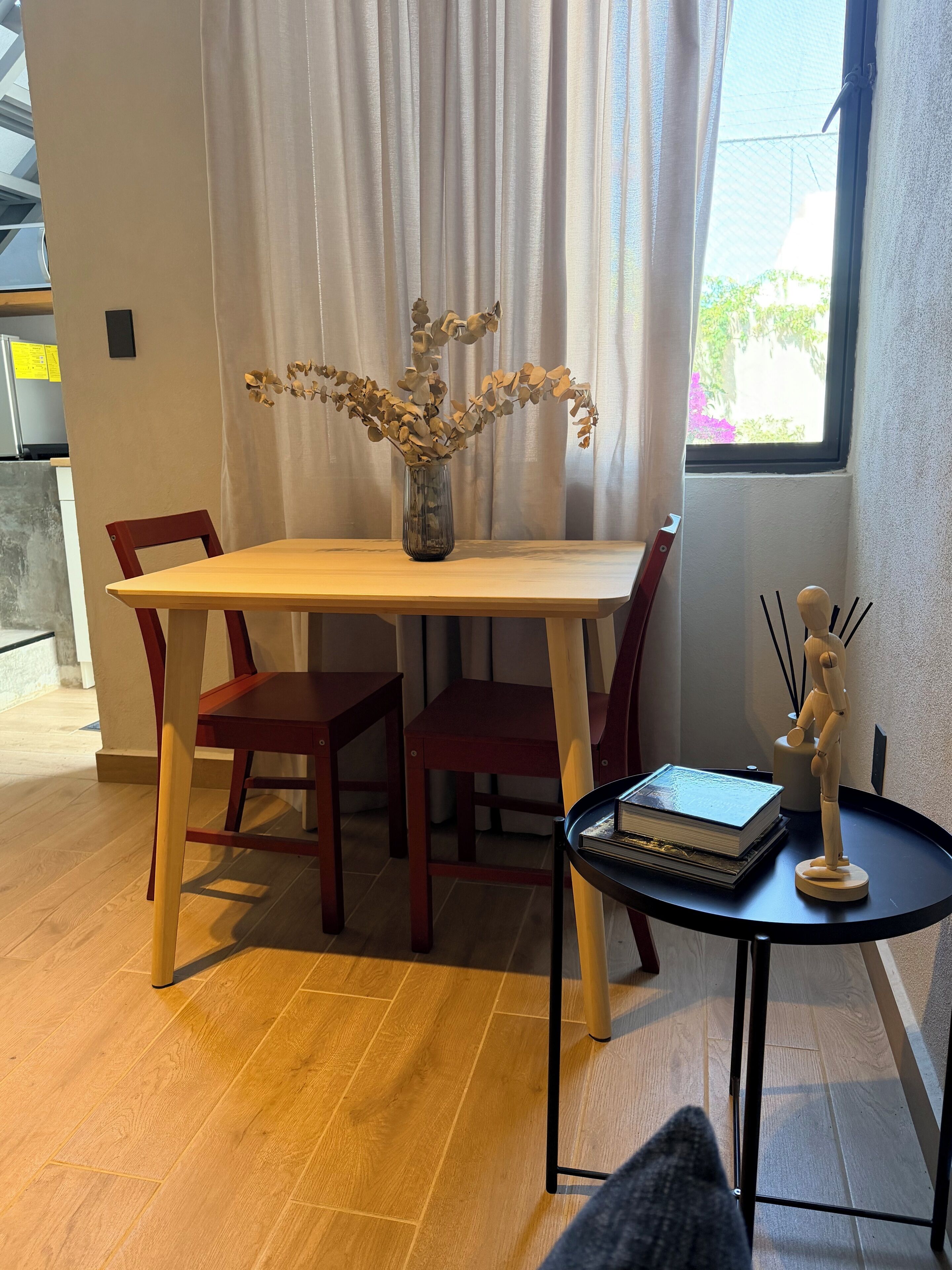 A dining area with a wooden table and chairs, a vase with dried flowers, and a small round table with a book and a wooden figure.
