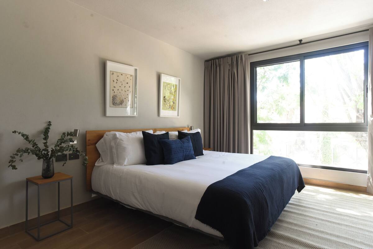 Un dormitorio con una cama, una mesita de noche, un florero con plantas y dos cuadros en la pared.