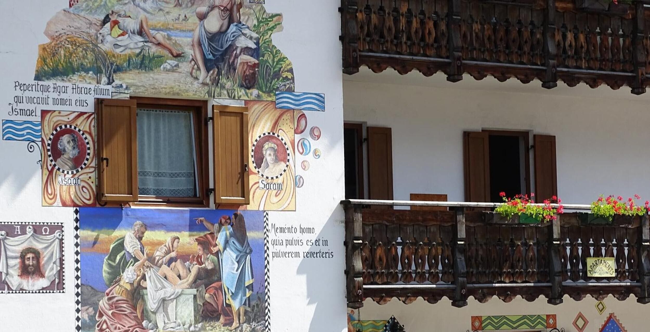 Un edificio con un balcone in legno, una finestra con tenda e un murale con figure religiose.