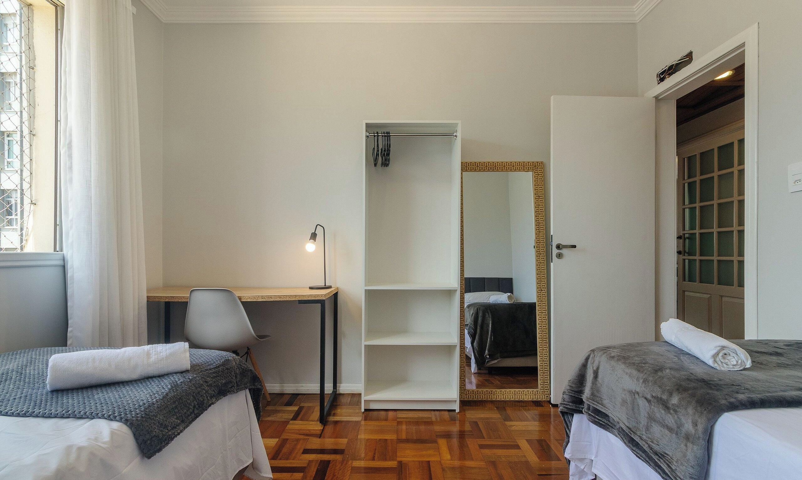 A hotel room with two beds, a desk, a chair, a wardrobe, and a mirror.