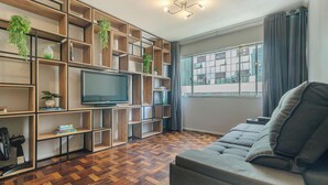 Ein Wohnzimmer mit einem großen Flachbildfernseher, Holzregalen mit Pflanzen, einem grauen Sofa und einem großen Fenster mit Blick auf die Stadt.