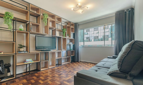 A living room with a television, wooden shelving, a grey sofa, and a window with curtains. - 3BR Fully Equipped apt in Floripa Dma0901 (Florianópolis)