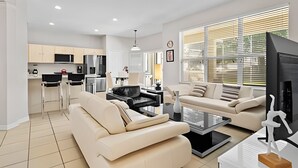 En moderne stue med sofa, sofabord og fjernsyn. Der er et køkkenområde med hårde hvidevarer og et spisebord.