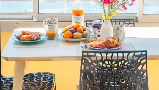 Mesa de desayuno con alimentos y un florero, con vista a un puerto deportivo con barcos.
