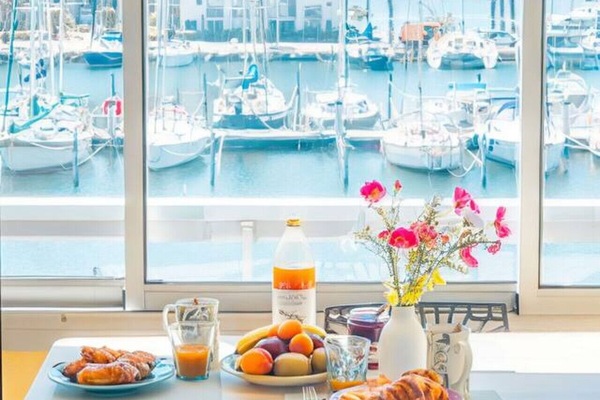 Petit-déjeuner avec vue sur le port de Leucate – table dressée avec fruits frais, jus d`orange et fleurs roses encadrées par la vue panoramique sur les voiliers. Un réveil de rêve au bord de l`eau.