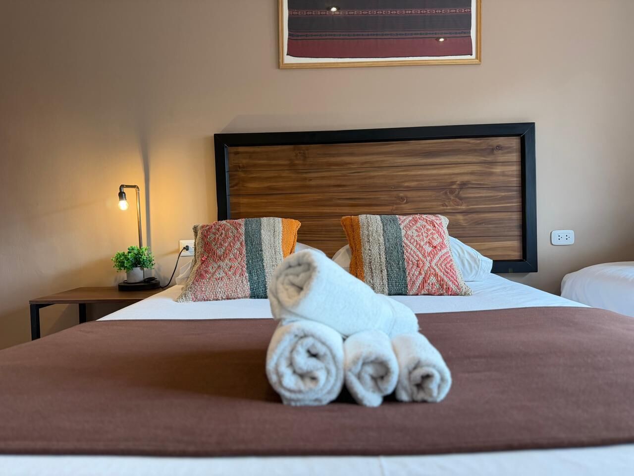A neatly made bed with rolled towels, a bedside lamp, a small plant, and patterned pillows.