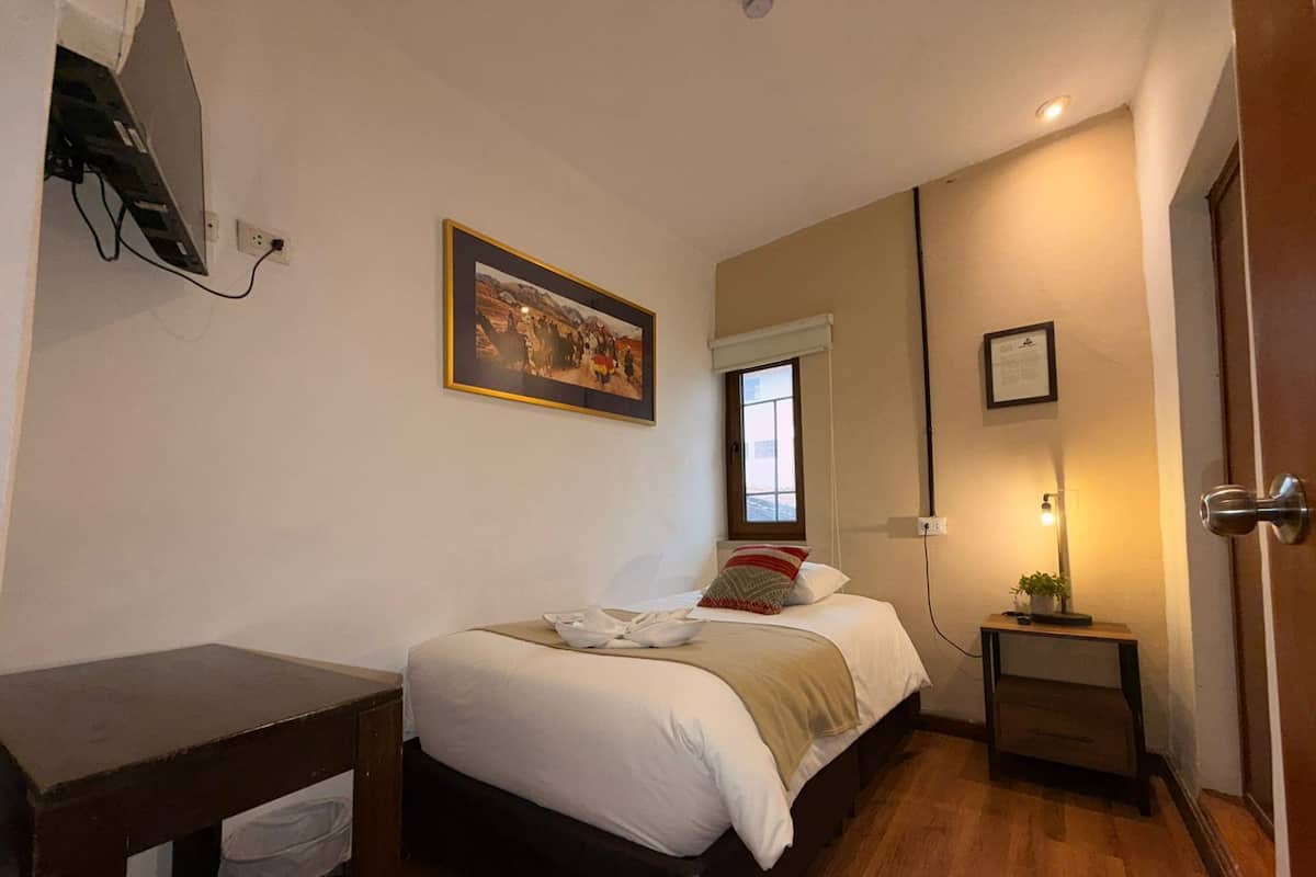Ein Schlafzimmer mit Bett, Nachttisch, kleinem Tisch mit Lampe, Fenster und einem Bild an der Wand.