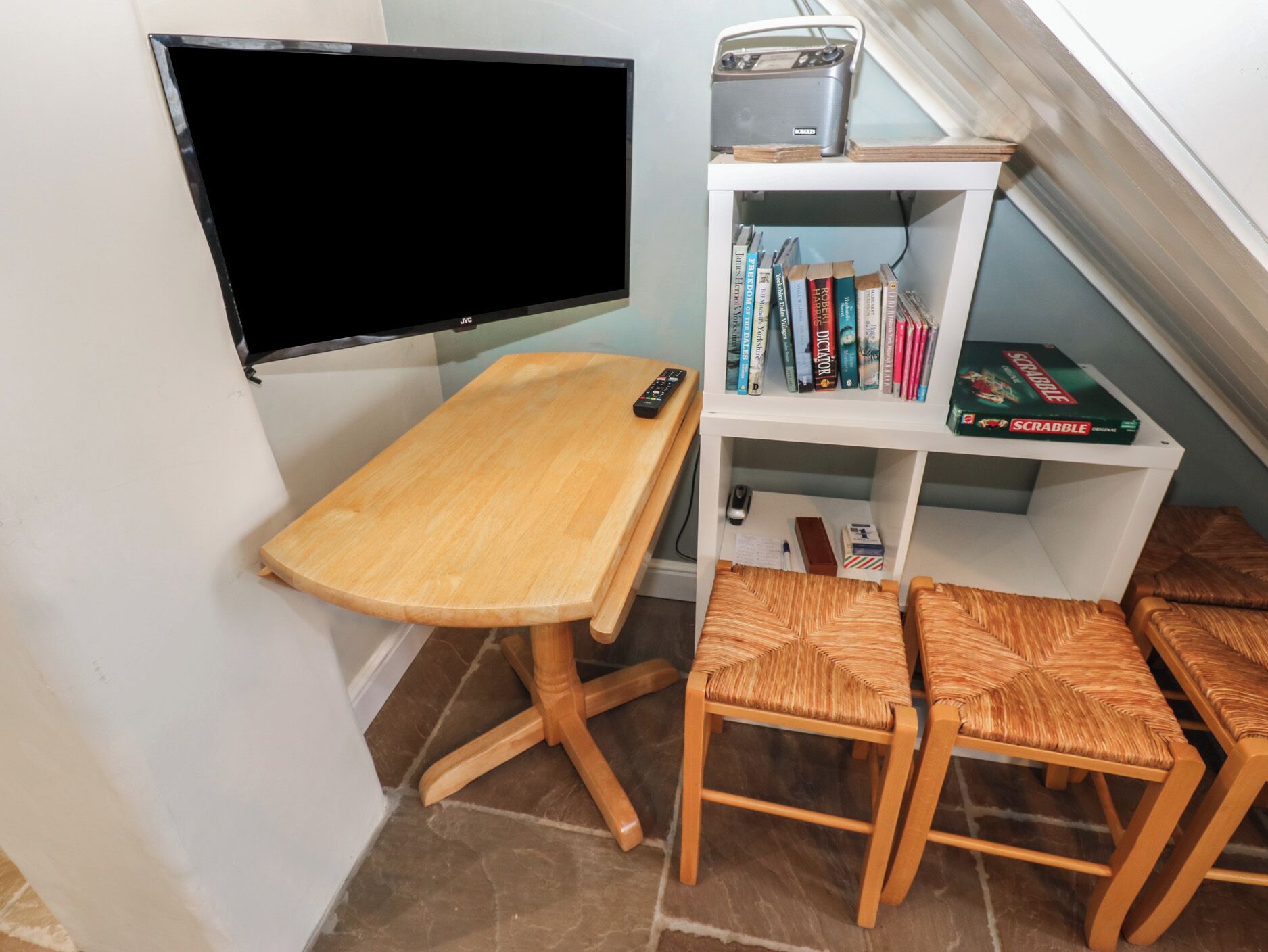 A small study area with a wooden desk, a television, a bookshelf with books and a Scrabble game, and two woven chairs.