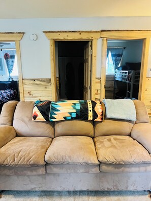 Ein beiges Ledersofa mit gemusterten Kissen und einer Decke, ein Fenster mit Holzrahmen und eine Tür, die in einen dunklen Raum führt.