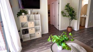 Ein modernes Wohnzimmer mit Flachbildfernseher, einer weißen Regaleinheit, einem runden weißen Hocker und einem Blumentopf.