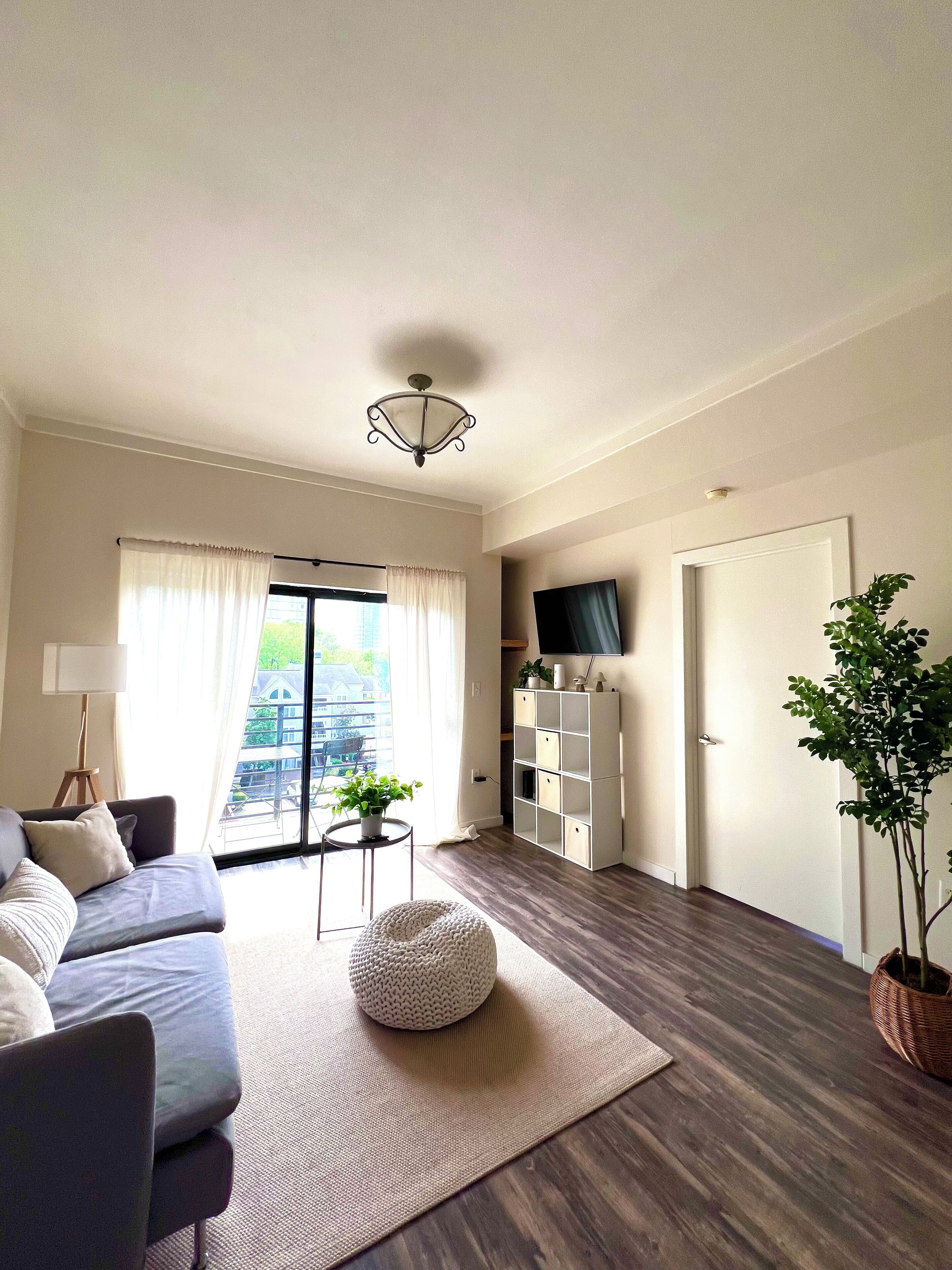 A modern living room with a grey sofa, a round white pouf, a small table with a plant, and a TV on a white shelf.