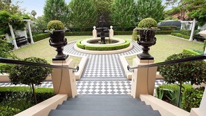 Un escalier menant à une fontaine circulaire entourée de verdure, avec un sol à carreaux et une pergola en arrière-plan.