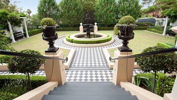 A staircase leading to a circular fountain surrounded by greenery, with a checkered floor and a pergola in the background.