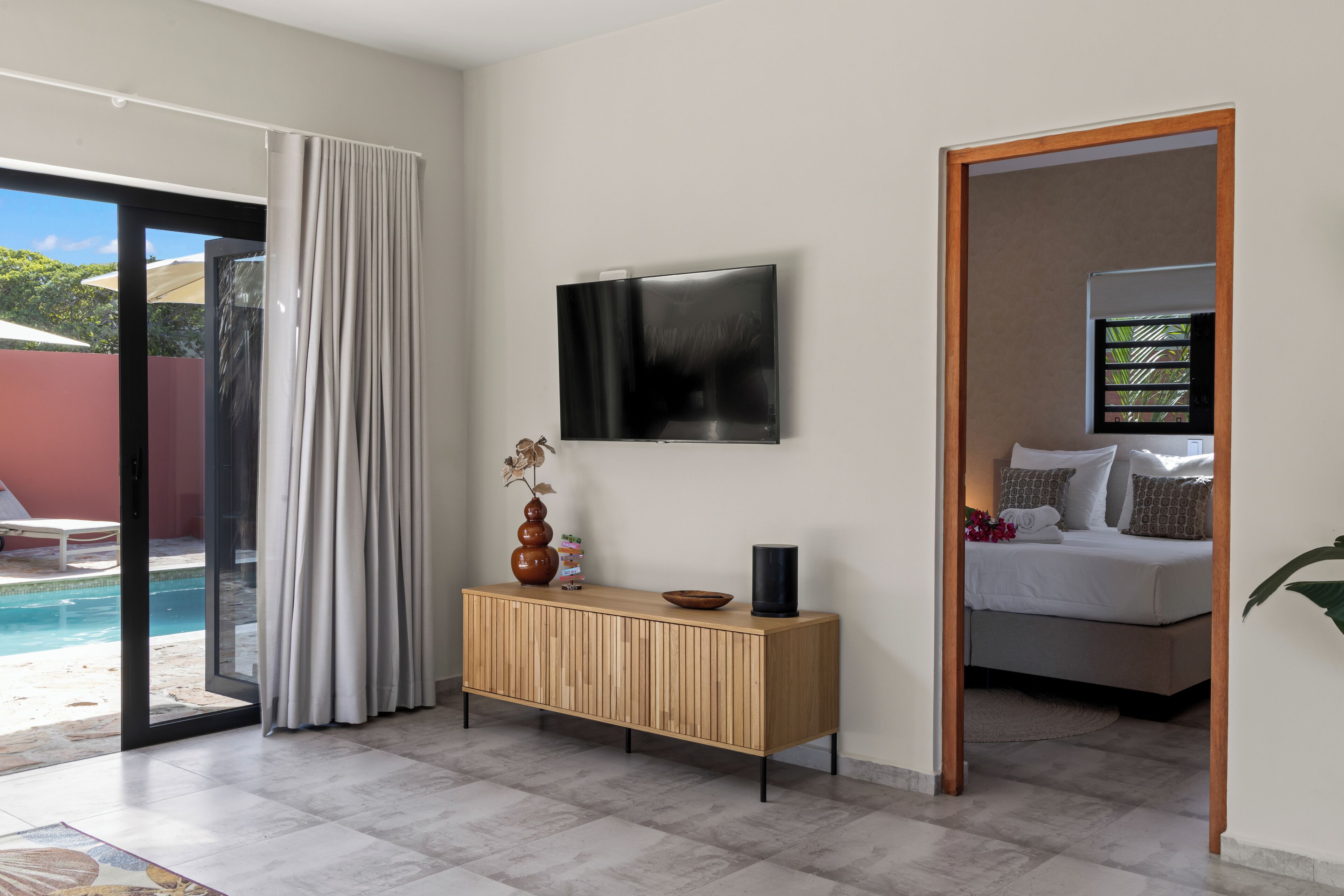 A modern hotel room with a large TV, a wooden console table, a bed with pillows, and a view of a swimming pool.