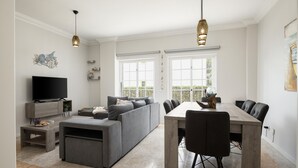 A modern living room with a grey sofa, a dining table with chairs, a TV, and pendant lights. - Mar e Golfe - Pool with golf view by HD (Vilamoura)