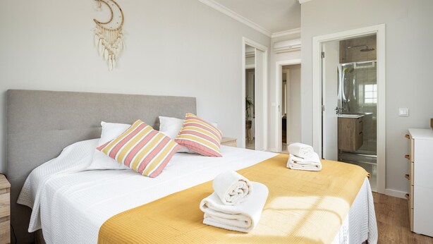 A neatly made bed with white bedding and a yellow throw, a bedside table with a lamp, and a wall-mounted dreamcatcher. - Mar e Golfe - Pool with golf view by HD (Vilamoura)