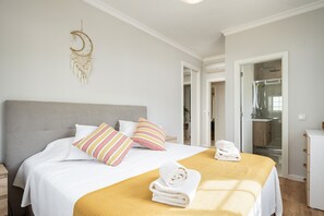 Ein ordentlich bezogenes Bett mit weißer Bettwäsche und einem gelben Überwurf, ein Nachttisch mit Lampe und ein an der Wand hängender Traumfänger.
