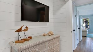 Une pièce dotée d’un téléviseur à écran plat fixé sur un mur recouvert de panneaux blancs, d’une console en osier agrémentée d’objets décoratifs et d’où l’on aperçoit une autre pièce par une porte ouverte.