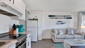 Une cuisine compacte avec des meubles blancs, une cuisinière et une machine à café. On y trouve un canapé beige agrémenté de coussins décoratifs et une petite table avec une lampe.