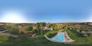 Ein großer Swimmingpool, umgeben von einer gepflegten Rasenfläche und Palmen, mit Blick auf entfernte Häuser und Felder.