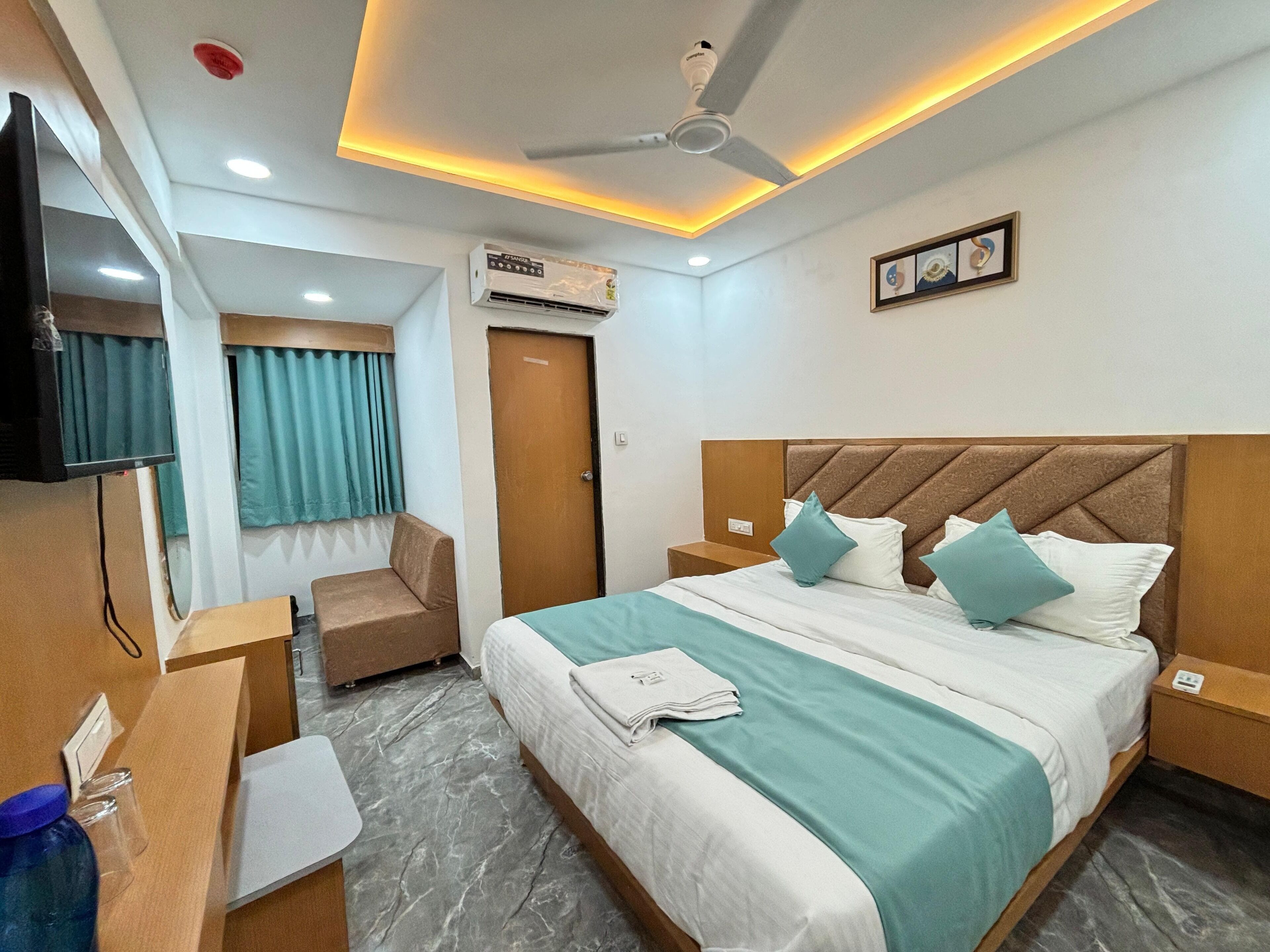 A hotel room with a bed, a desk, a chair, a television, an air conditioning unit, and a ceiling fan.