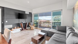A modern living room with a large sofa, a coffee table, and a flat-screen TV mounted on the wall. There is a balcony with a view of a cruise ship and buildings. - Deck 12 Vallarta Residences (Puerto Vallarta)