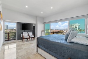 Una camera da letto moderna con un letto grande, una TV a schermo piatto montata sulla parete, due poltrone e un balcone con vista sulla città.