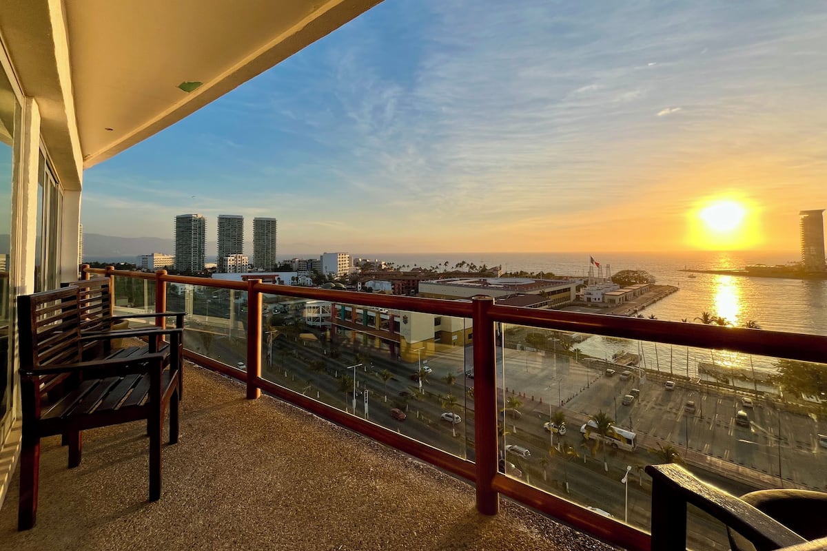 A balcony with a view of a sunset over a body of water, buildings, and a road.