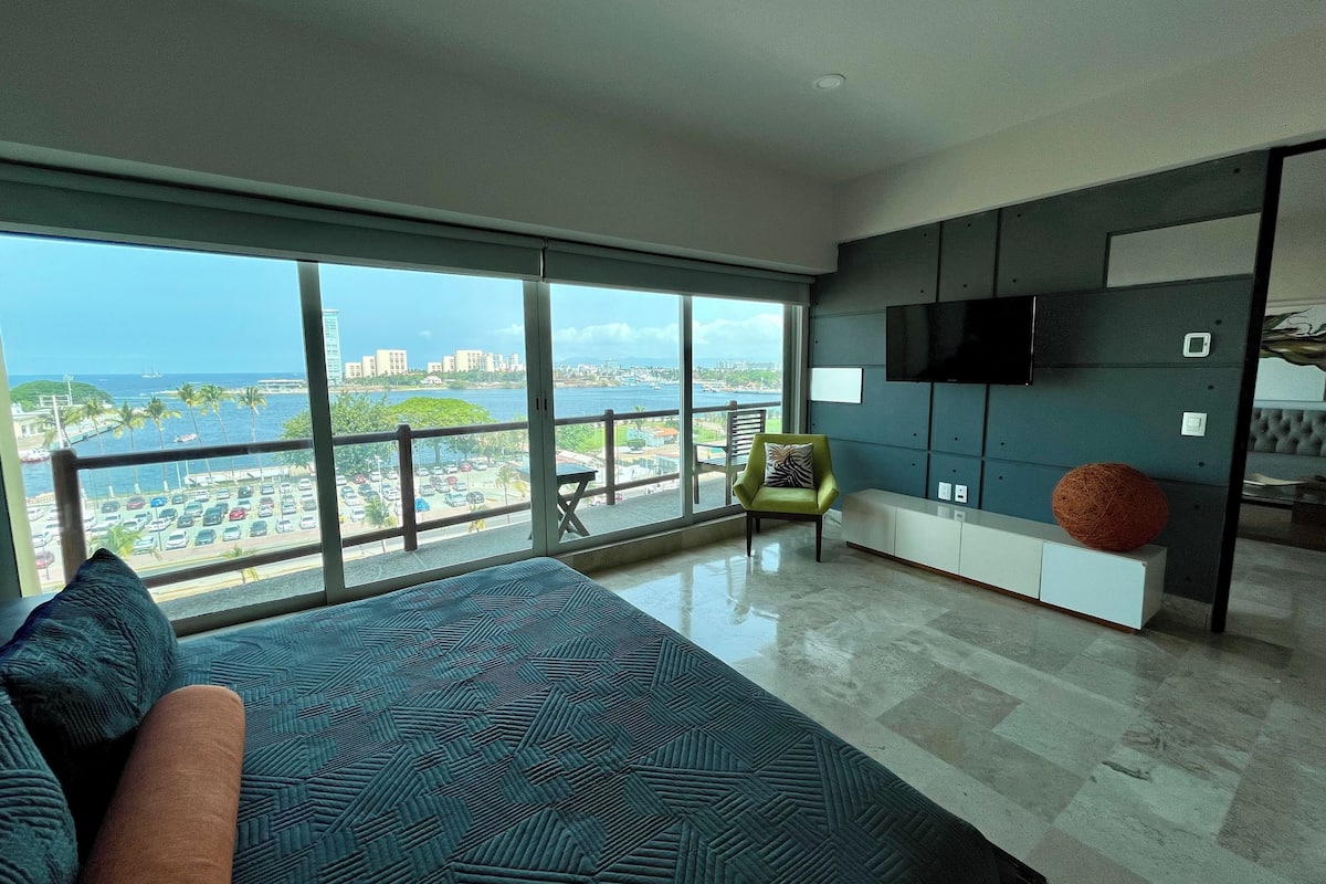 A modern bedroom with a large window, a bed, a green chair, and a TV on the wall.