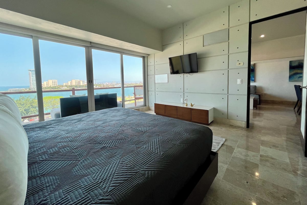 A modern bedroom with a large bed, a TV mounted on the wall, and a view of the city and water.