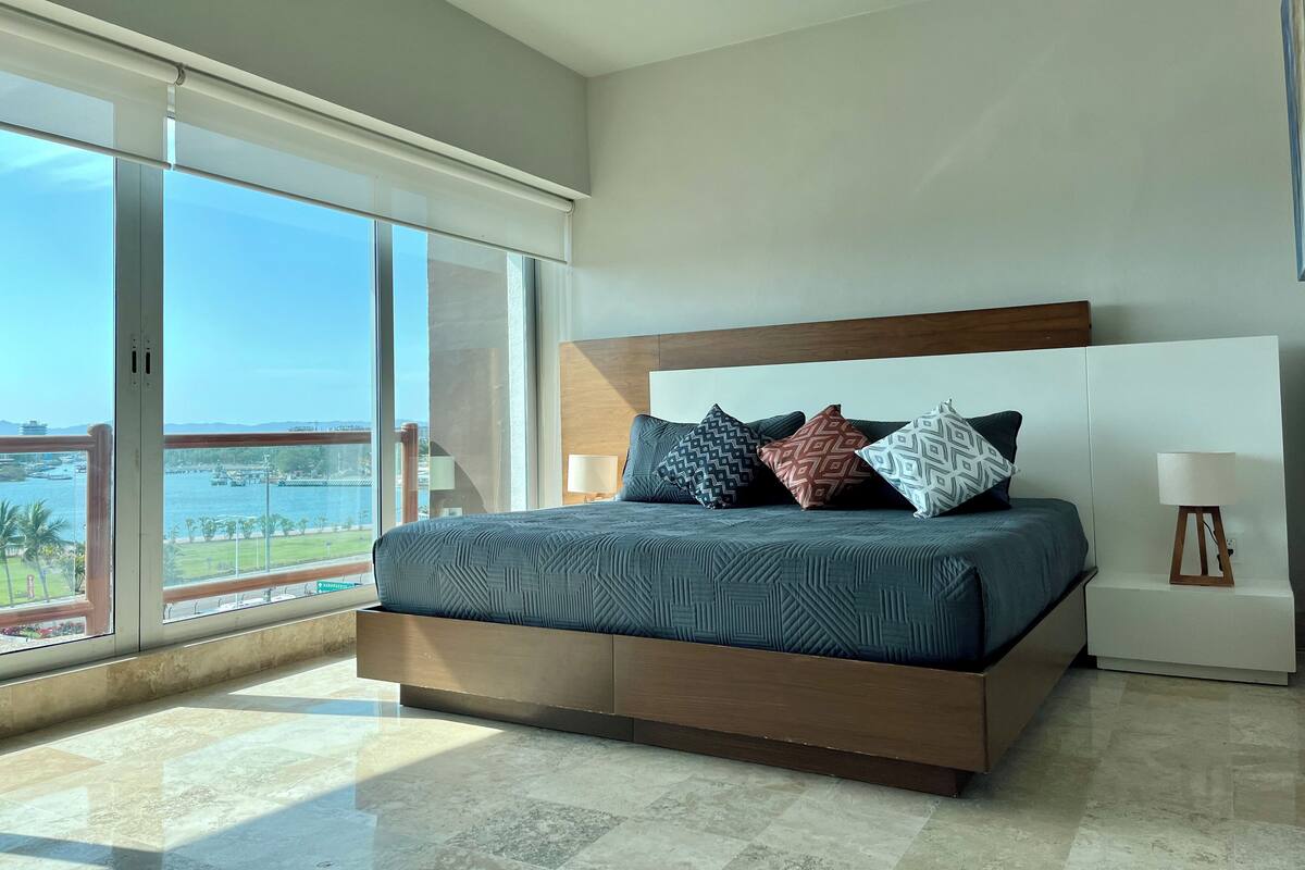 A modern bedroom with a large bed, a wooden headboard, and a view of the sea through large windows.