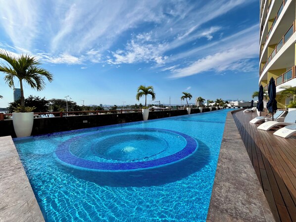 Una piscina con bordo in legno e poltroncine, circondata da palme e cielo sereno.