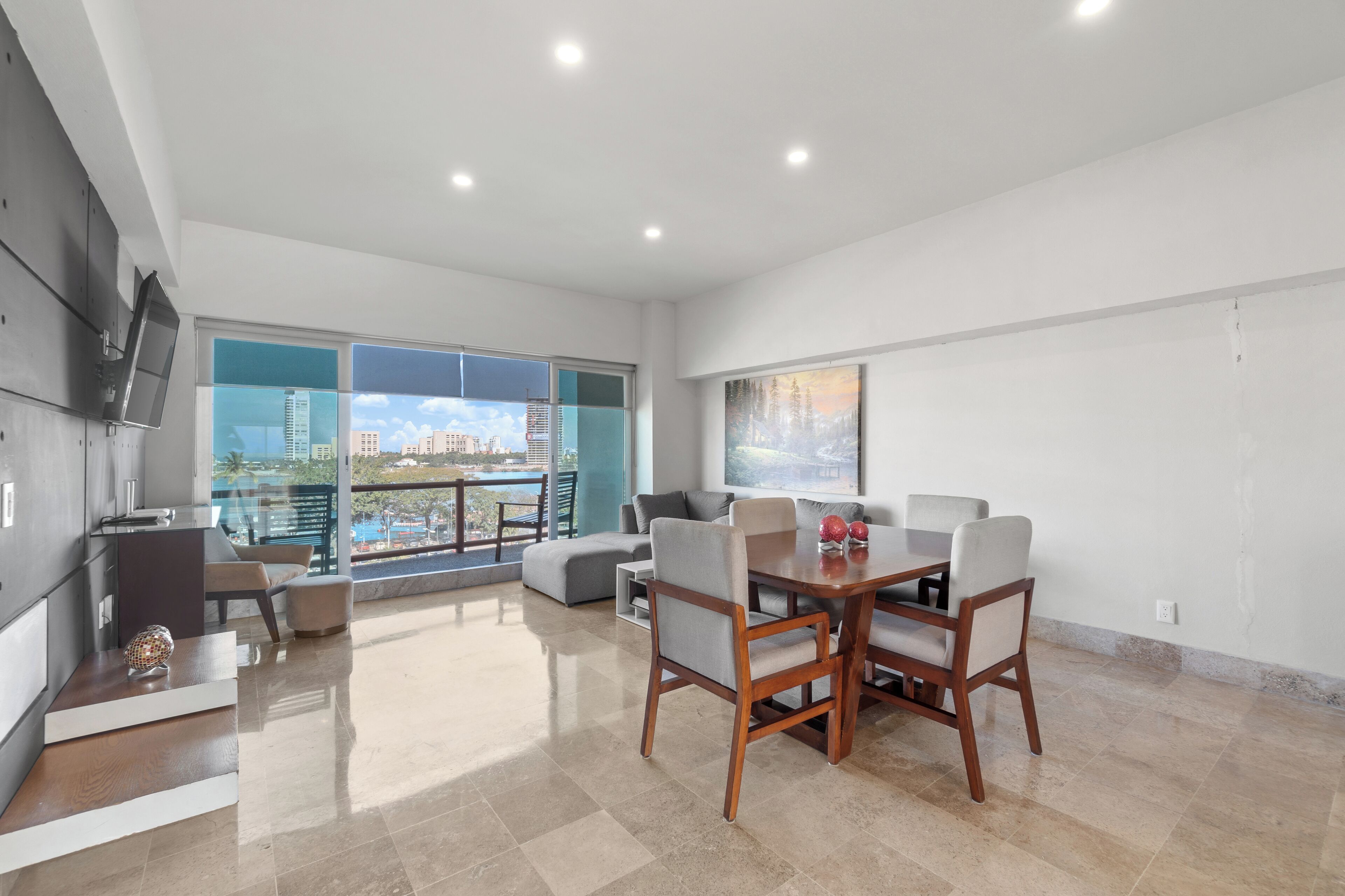 A spacious living area with a dining table, chairs, and a sofa. There is a TV mounted on the wall and a balcony with a view of the city.