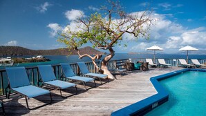 Una terraza de madera con sillones de descanso y piscina, con vistas al mar azul cristalino y colinas al lejano.
