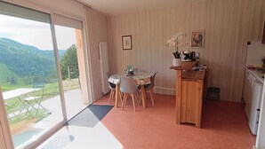 Une pièce comprenant une table à manger, des chaises, un petit coin cuisine et une vue sur un paysage verdoyant, visible à travers une grande fenêtre.