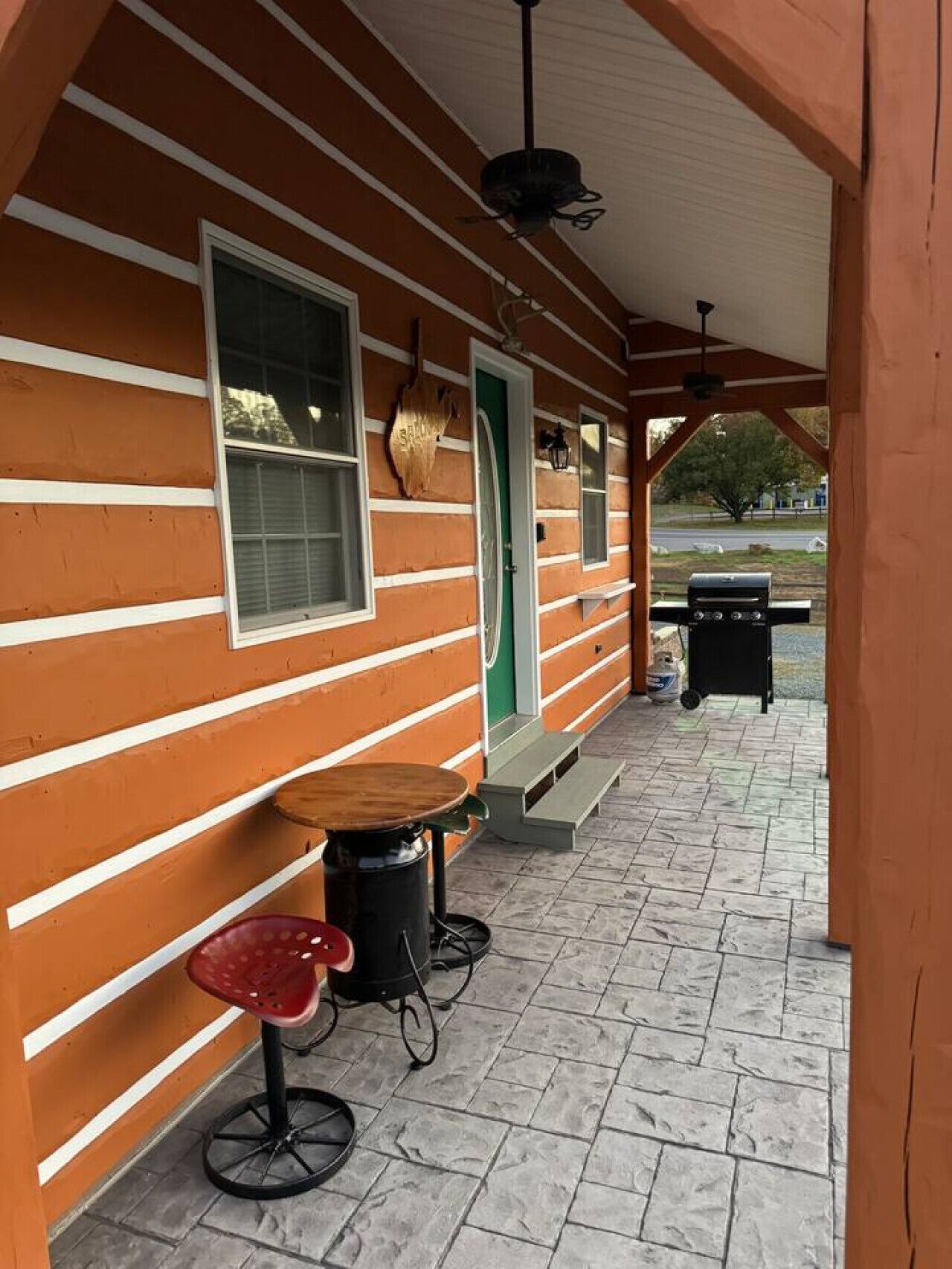 A covered porch with a table and a red stool, a grill, and a green door.