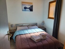 Una cama bien hecha con una colcha rosa y azul, dos almohadas y dos toallas dobladas sobre una mesita de noche de madera con una lámpara.