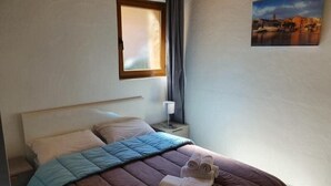 A neatly made bed with a brown and blue quilt, a nightstand with a lamp, and a painting of a cityscape on the wall. - casa del torrent (Ota)