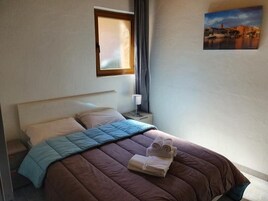 Una cama bien hecha con colcha marrón y azul, una mesita de noche con lámpara, y un cuadro con una vista urbana en la pared.
