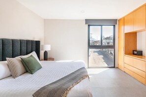 A bedroom with a large bed, a nightstand, a lamp, and a view of the city through a sliding door. - Cozy Downtown Apartment Perfect for Couples (Puerto Vallarta)