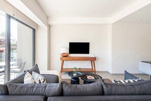 A modern living room with a sofa, a coffee table, a TV on a wooden stand, and a view through large windows. - Cozy Downtown Apartment Perfect for Couples (Puerto Vallarta)