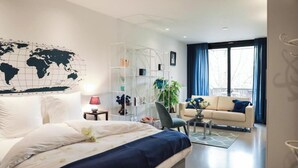 A modern hotel room with a large bed, a seating area with a sofa and armchairs, a world map on the wall, and a balcony with curtains. - Suite aux Anges N°1 (Antwerpen)