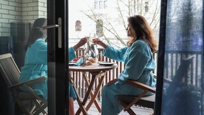 Two people in matching teal bathrobes seated at a round table on a balcony with a view of buildings and trees. - Suite aux Anges N°1 (Antwerpen)