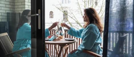 Two people in matching teal bathrobes seated at a round table on a balcony with a view of buildings and trees.