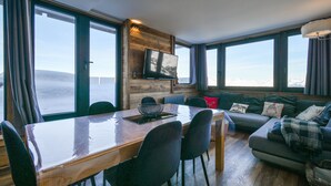 A modern living room with a wooden dining table, black chairs, a sofa, and a TV mounted on a wooden wall. - Apartment “Lesetoilesdelaplagne - Le Diamant” with Mountain View and Private Terrace (aime la plagne)