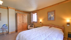 A bedroom with a large bed, wooden wardrobe, chair, and a painting on the wall.