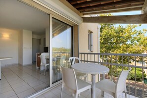 A balcony with a table and chairs, a sliding glass door, and a view of trees and buildings. - Apartment 'Cap d’Agde - Primavera - 1' with Shared Pool, Private Terrace and Air Conditioning (Agde)
