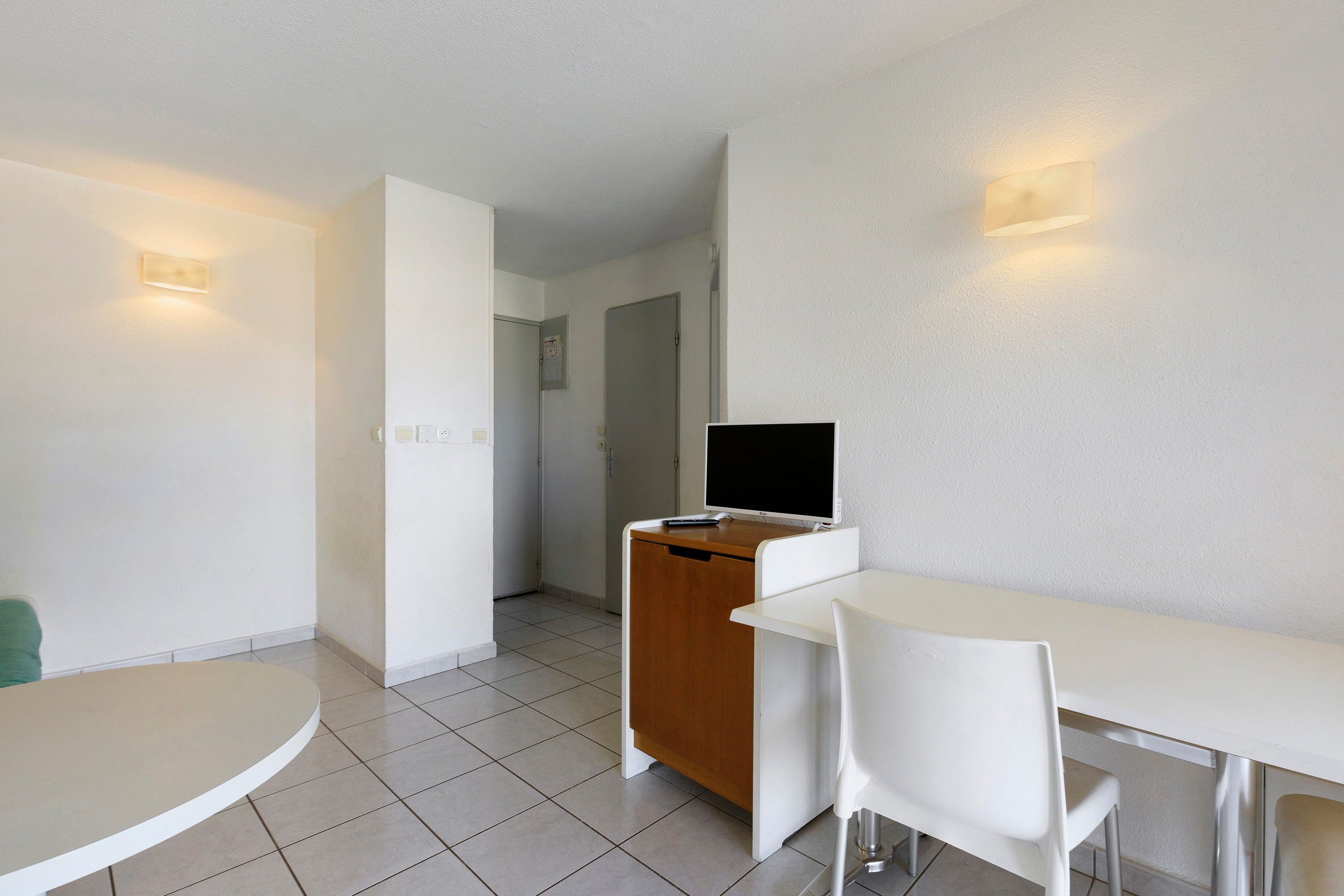 Ein Hotelzimmer mit einem weißen Schreibtisch, einem Sessel, einem Fernseher auf einer Holzkommode und einer Tür, die in einen anderen Raum führt.