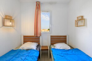 A room with two beds, each with a wooden headboard, white pillows, and blue bedding. There is a window with a view of buildings and a striped curtain. - Apartment 'Cap d’Agde - Primavera - 1' with Shared Pool, Private Terrace and Air Conditioning (Agde)