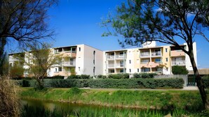 A residential building with multiple balconies, a pond, and surrounding greenery. - Apartment 'Cap d'Agde - Primavera - 2' with Sea View, Shared Pool and Air Conditioning (Agde)